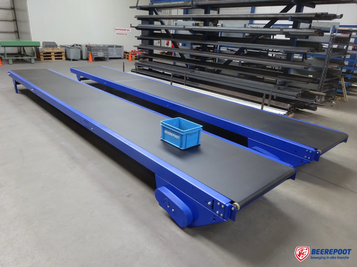 Je ziet twee blauwe transportbanden van Beerepoot netjes naast elkaar en even groot in een voorraadhal van Beerepoot. Op de voorste band staat een blauw kratje van Beerepoot. Op de achtergrond zie je voorraad van Beerepoot.