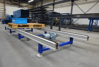 Op de foto staat een kettingbaan met blauwe pootondersteuningen. Het frame is aluminium. De aandrijving is lichtblauw van het merk SEW. De kettingbaan staat opgesteld in de werkplaats van Beerepoot BV.