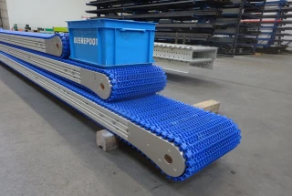 Je ziet drie blauwe schakelbanden van Beerepoot op elkaar gestapeld met daarop een blauw Beerepoot kratje. De schakelbanden staan in de werkplaats van Beerepoot. Op de achtergrond staat nog een machine en kasten met onderdelen.