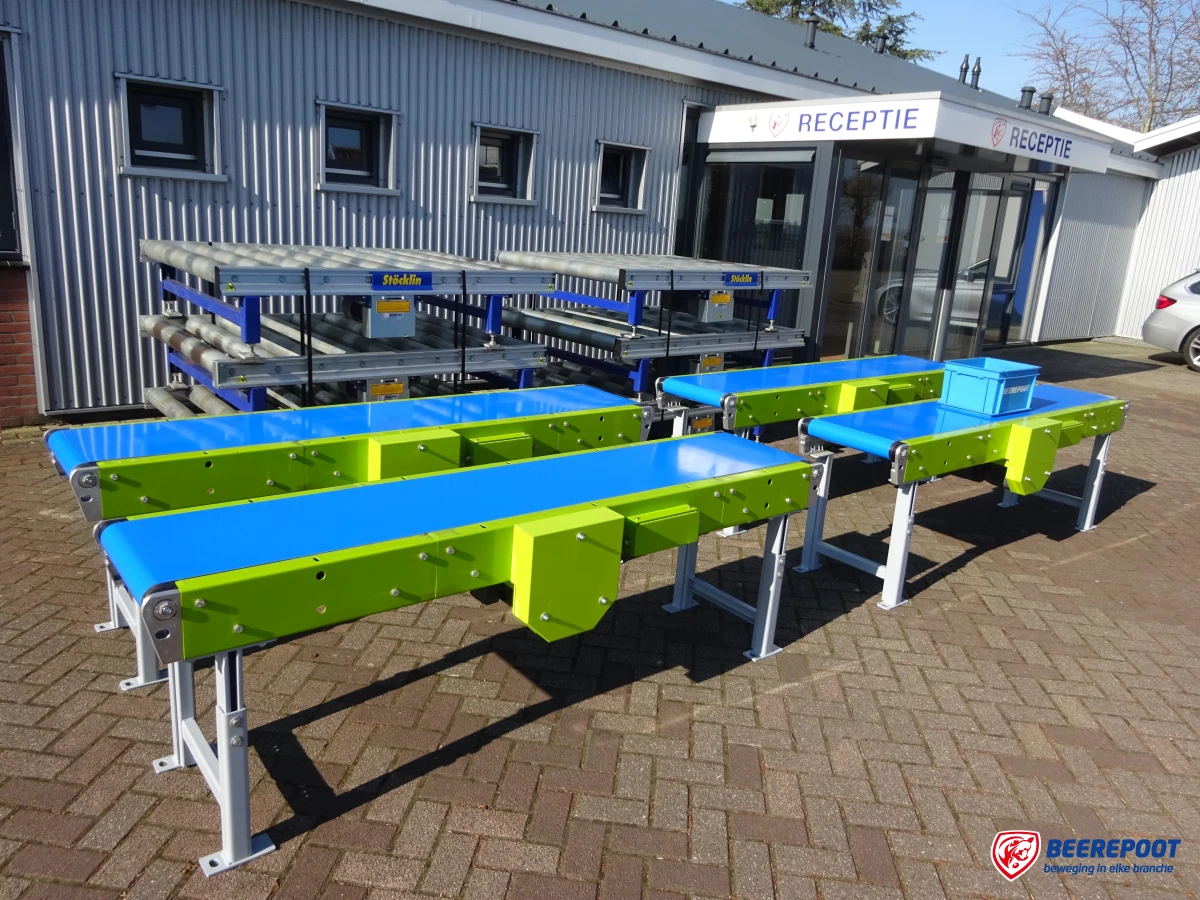 Je ziet vier blauwe en groene vlakke transportbanden van Beerepoot, twee naast elkaar en twee voor elkaar. Erachter staat nog een aantal kleine rollenbanen (grijs met blauw). De banden staan voor het pand van Beerepoot uitgestald.