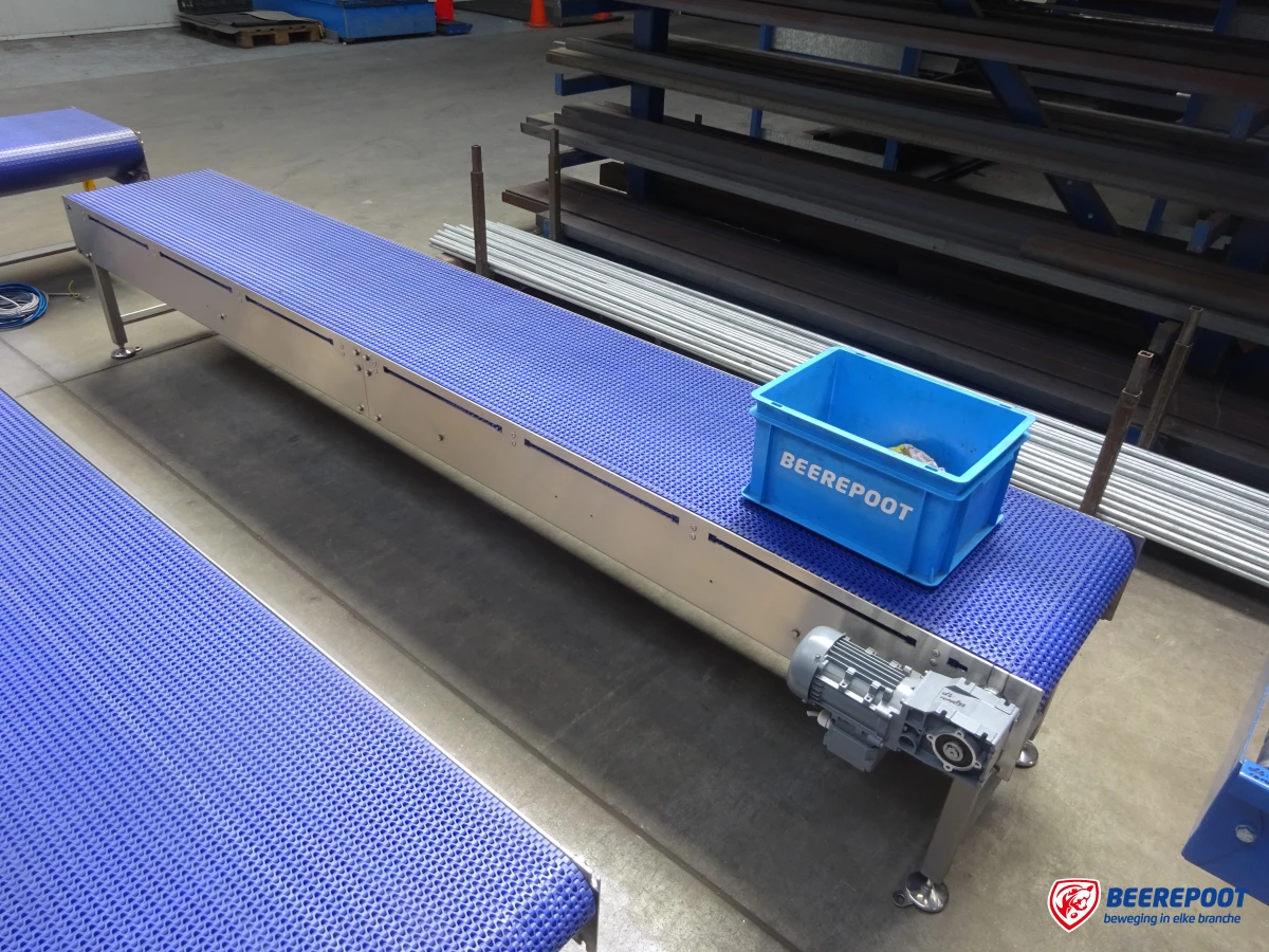 Je ziet een blauwe schakelband van Beerepoot met daarop een blauw Beerepoot kratje in de werkplaats van Beerepoot. Ervoor en links staat nog een blauwe schakelband. Op de achtergrond staat een rek met diverse onderdelen.