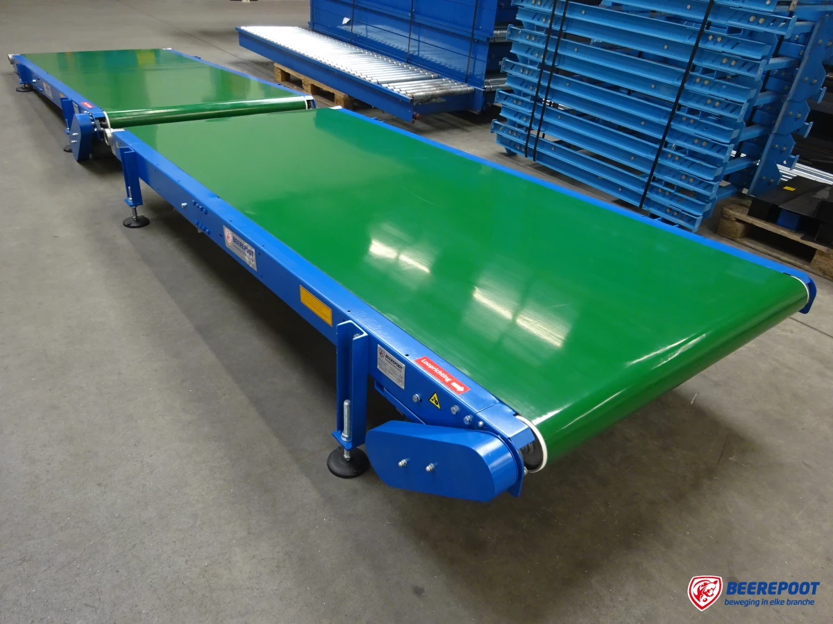 Je ziet twee groen/ blauwe vlakke bandtransporteurs van Beerepoot achter elkaar staan in een voorraadhal van Beerepoot. Op de achtergrond staan diverse korte blauwe bandtransporteurs op elkaar gestapeld.