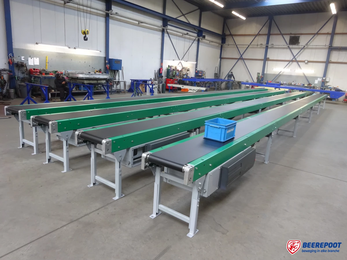Je ziet vier lange groene vlakke bandtransporteurs van Beerepoot uitgestald in de werkplaats van Beerepoot. Je ziet een blauw kratje van Beerepoot op de voorste bandtransporteur.
