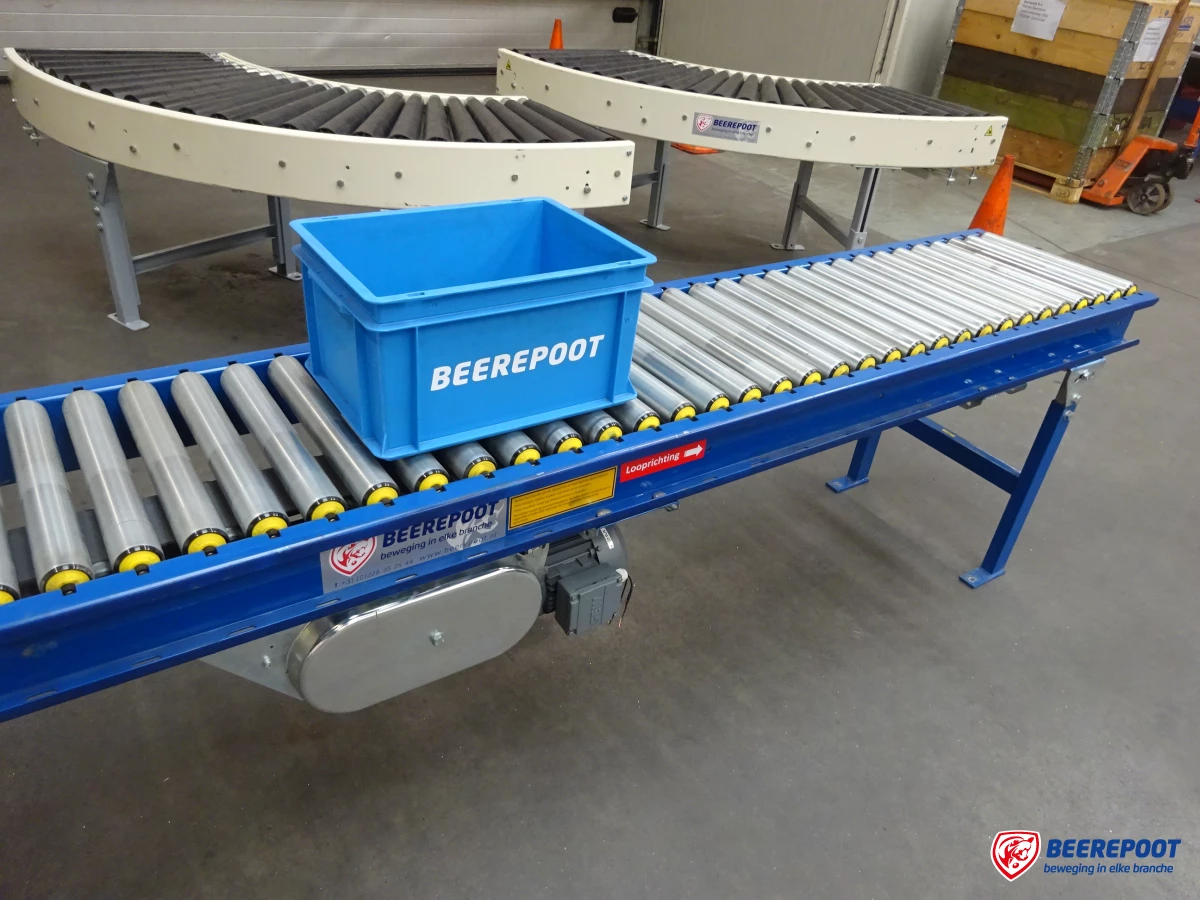 Je ziet een aangedreven blauwe rollenbaan met daarop een blauw Beerepoot kratje. Op de achtergrond zie je ook een aantal rollenbaanbochten. Het is een beeld uit een vooraadhal van Beerepoot